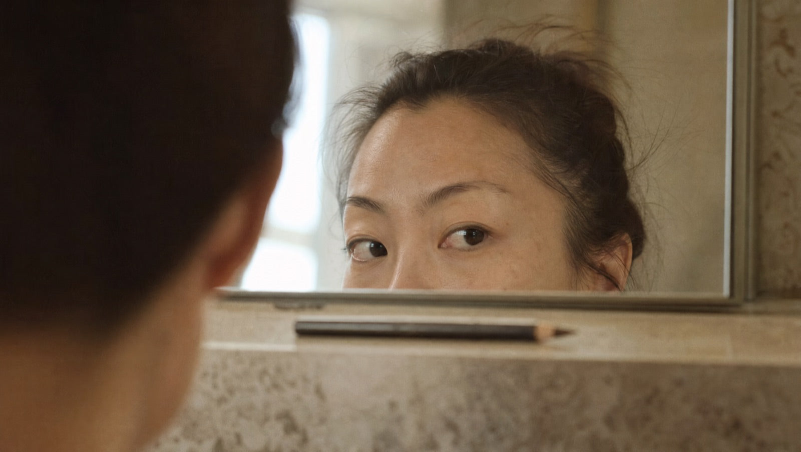 Close mirror reflection — the outer third of the brows sparse and faded toward the temples, a brow pencil resting on the counter