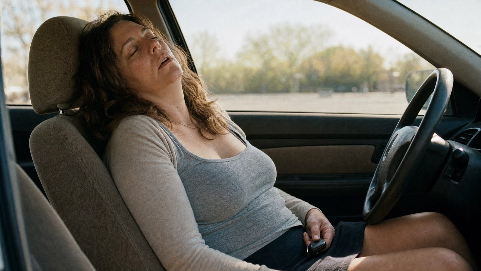 A woman slumped asleep in the driver's seat of a parked car in mid-afternoon light, keys still in her hand