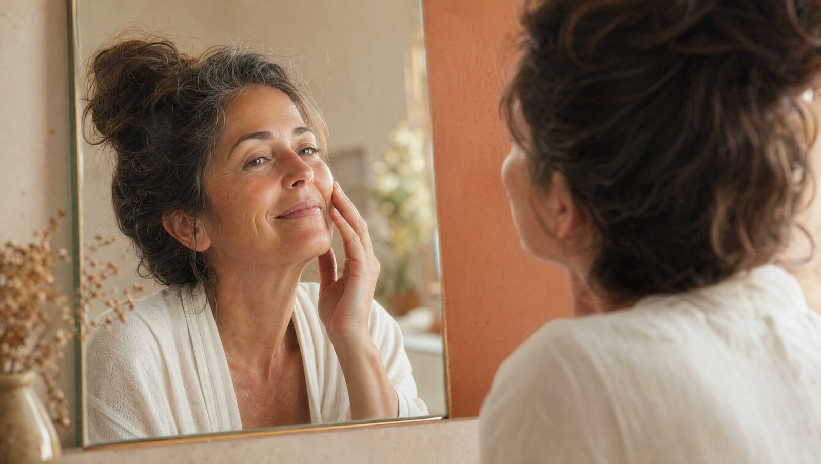 A woman at ease with her own face, looking at her reflection with quiet contentment in warm morning light