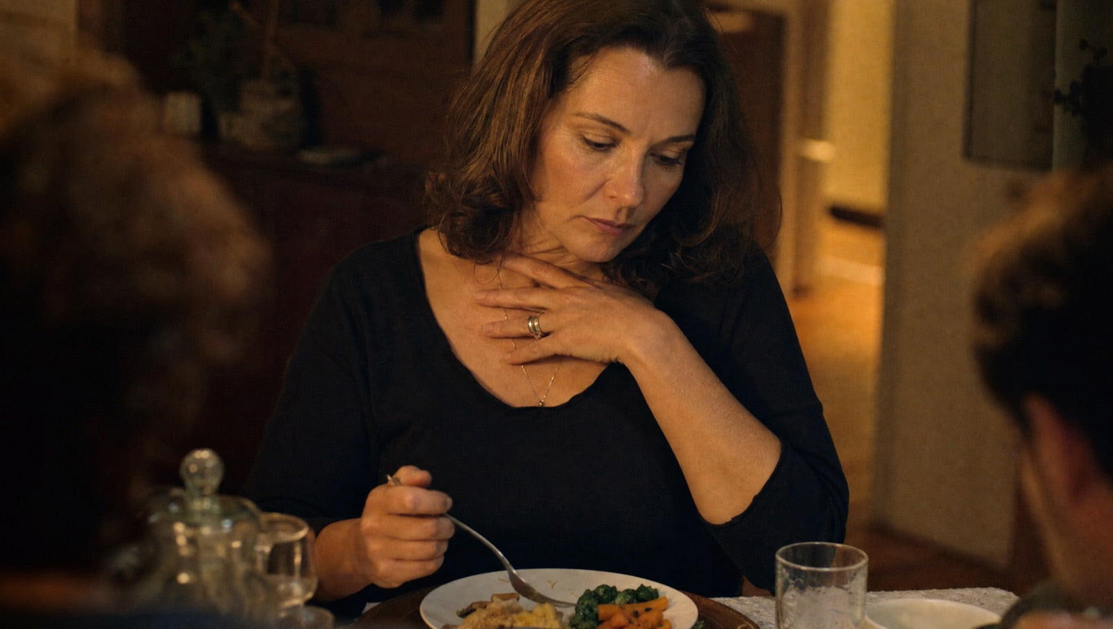 A woman eating slowly and carefully at a table, a hand near the base of her throat