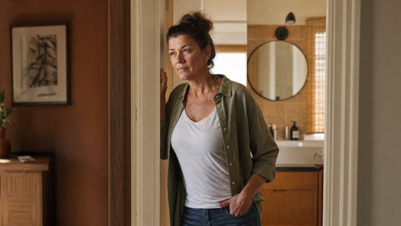 A woman pauses in the doorway of her bathroom in mid-afternoon light, tired and watchful